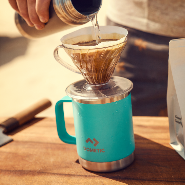 Person pouring coffee into a Dometic cup on a wooden surface.