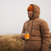 Person wearing a brown puffer jacket holding an orange mug in a natural setting