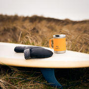 Orange Dometic mug and black mittens on a surfboard with grassy background