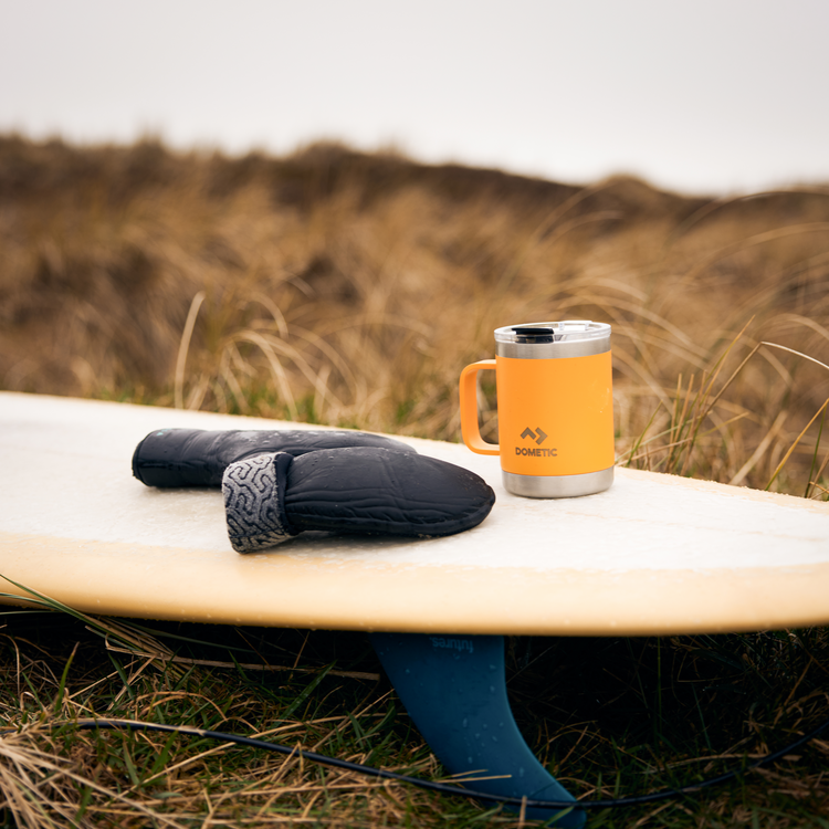 Orange Dometic mug and black mittens on a surfboard with grassy background