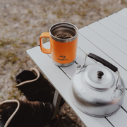 Orange Dometic mug with metal lid and silver kettle on a wooden surface outdoors.