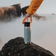 Person holding a blue insulated bottle on a rock with a blurred natural background