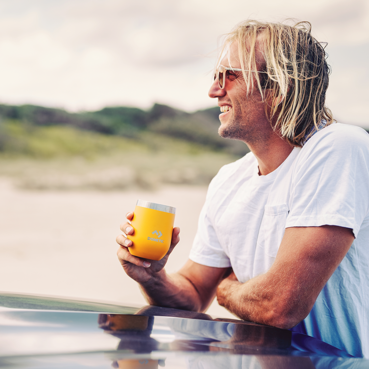 Man holding a yellow mug by a lake with mountains in the background