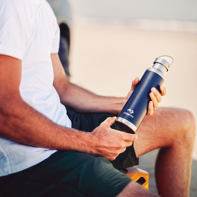 Person holding a dark gray water bottle with a silver cap, sitting outdoors.