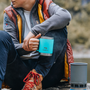 Person in outdoor gear holding a blue Dometic mug with a blurred natural background