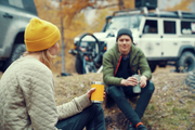 Two people sitting outdoors with yellow cups, surrounded by autumn foliage and vehicles.
