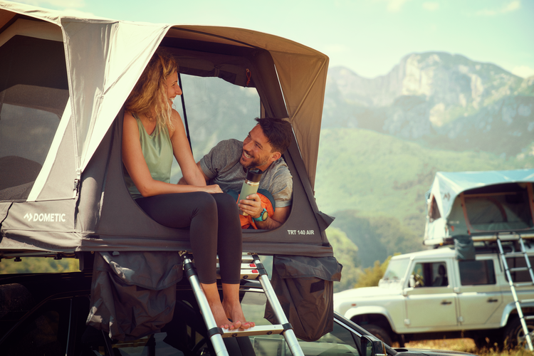 Two people sitting in a rooftop tent with mountains in the background