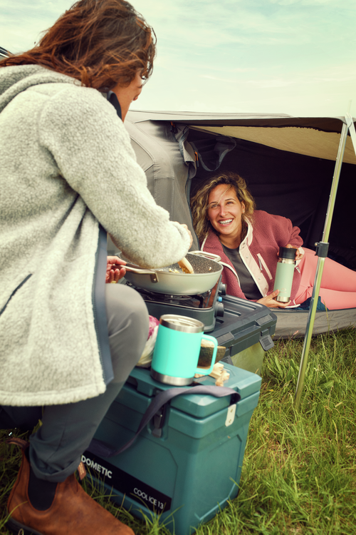 Two people camping outdoors, one cooking by a cooler with a pot and the other sitting inside a tent.