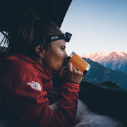 Person in a red jacket drinking from an orange mug with mountains in the background