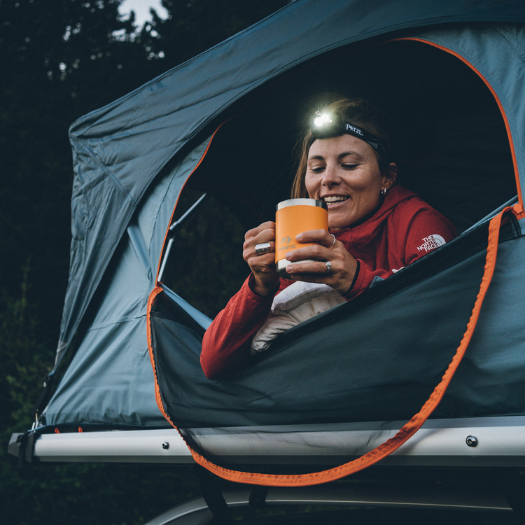 Person inside a tent holding a mug with a headlamp, sitting on a vehicle at night.