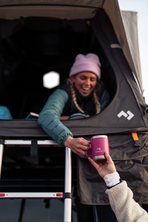 Person handing a pink Dometic mug to another person from inside a tent