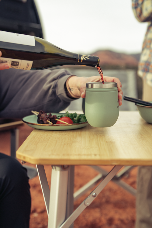 Person pouring red wine into a green cup at a table with food and scenic background