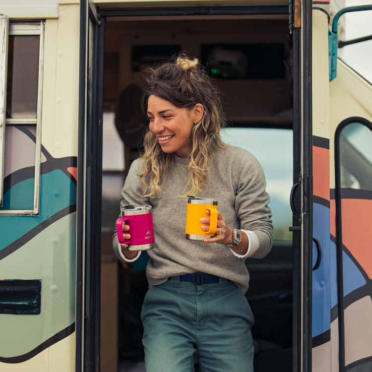 Woman standing in the doorway of a colorful vehicle holding two mugs.