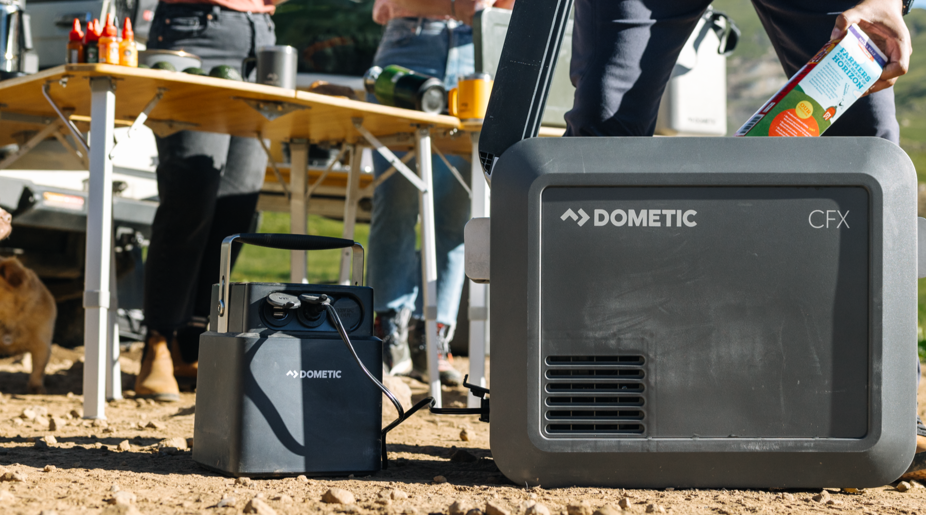 Dometic CFX5 electric cooler on a dirt ground with people and outdoor furniture in the background.