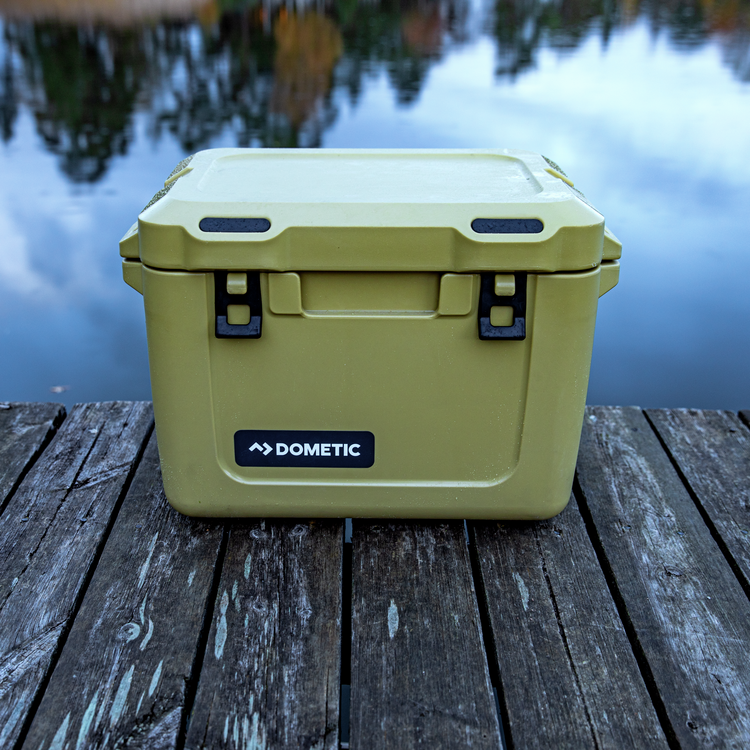 Olive colored Dometic cooler on a wooden surface by a body of water.
