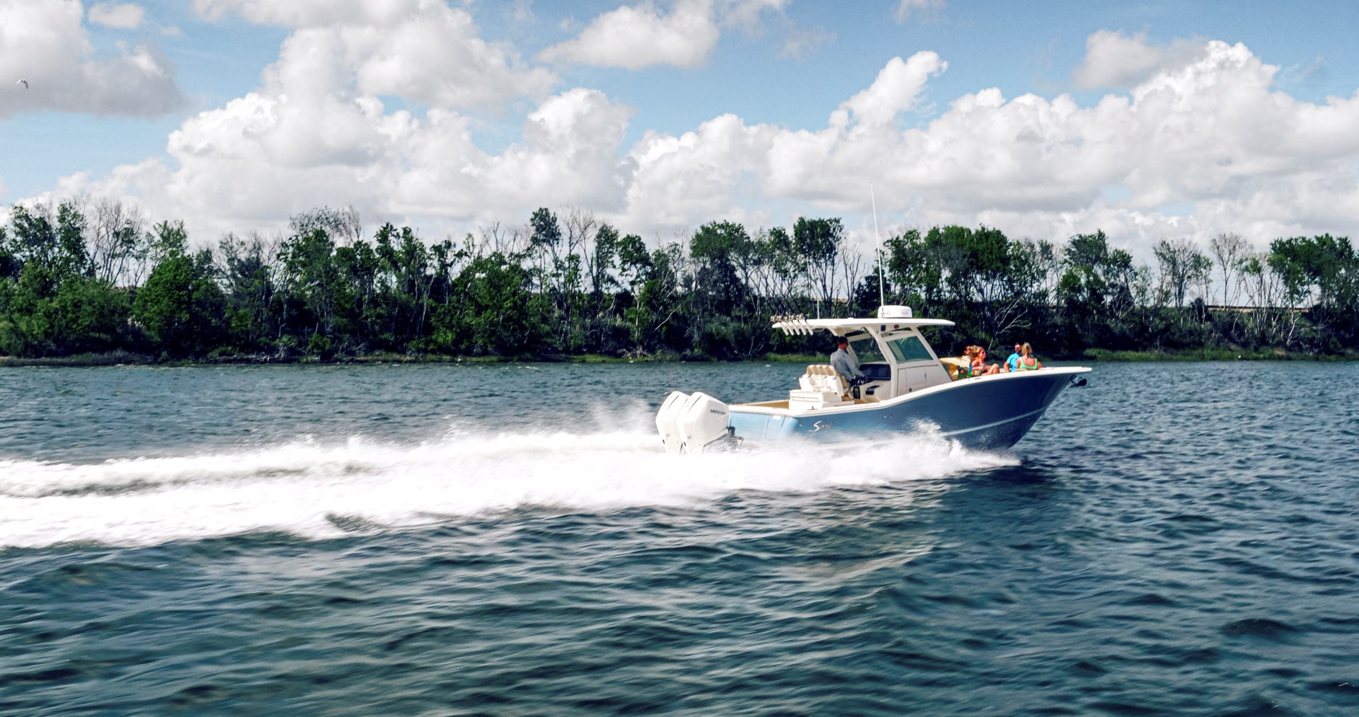 A center console fishing boat moving at a high speed along a tree-filled shoreline