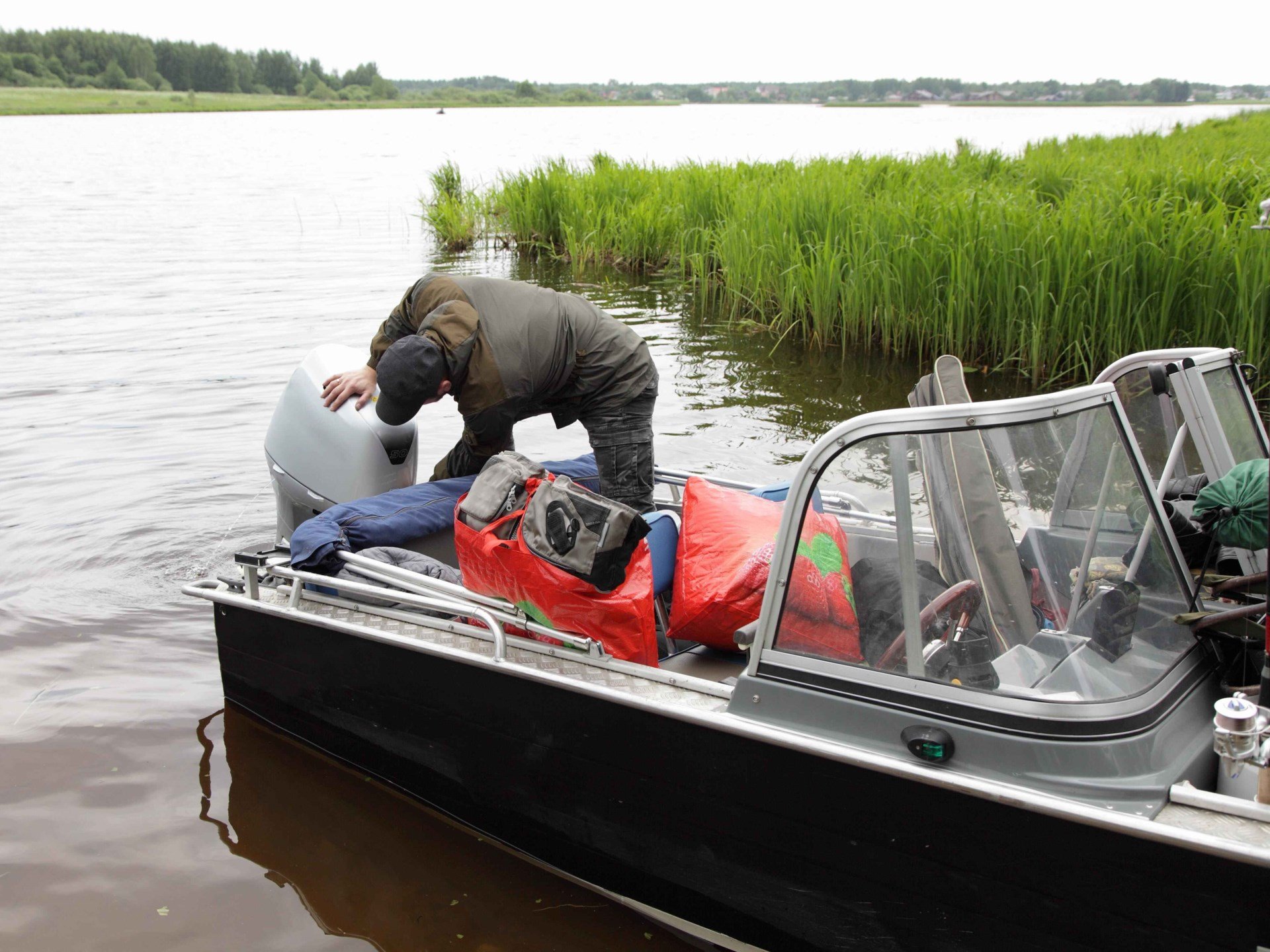 A man attempting a boat repair while sitting in calm water surrounded by green grass
